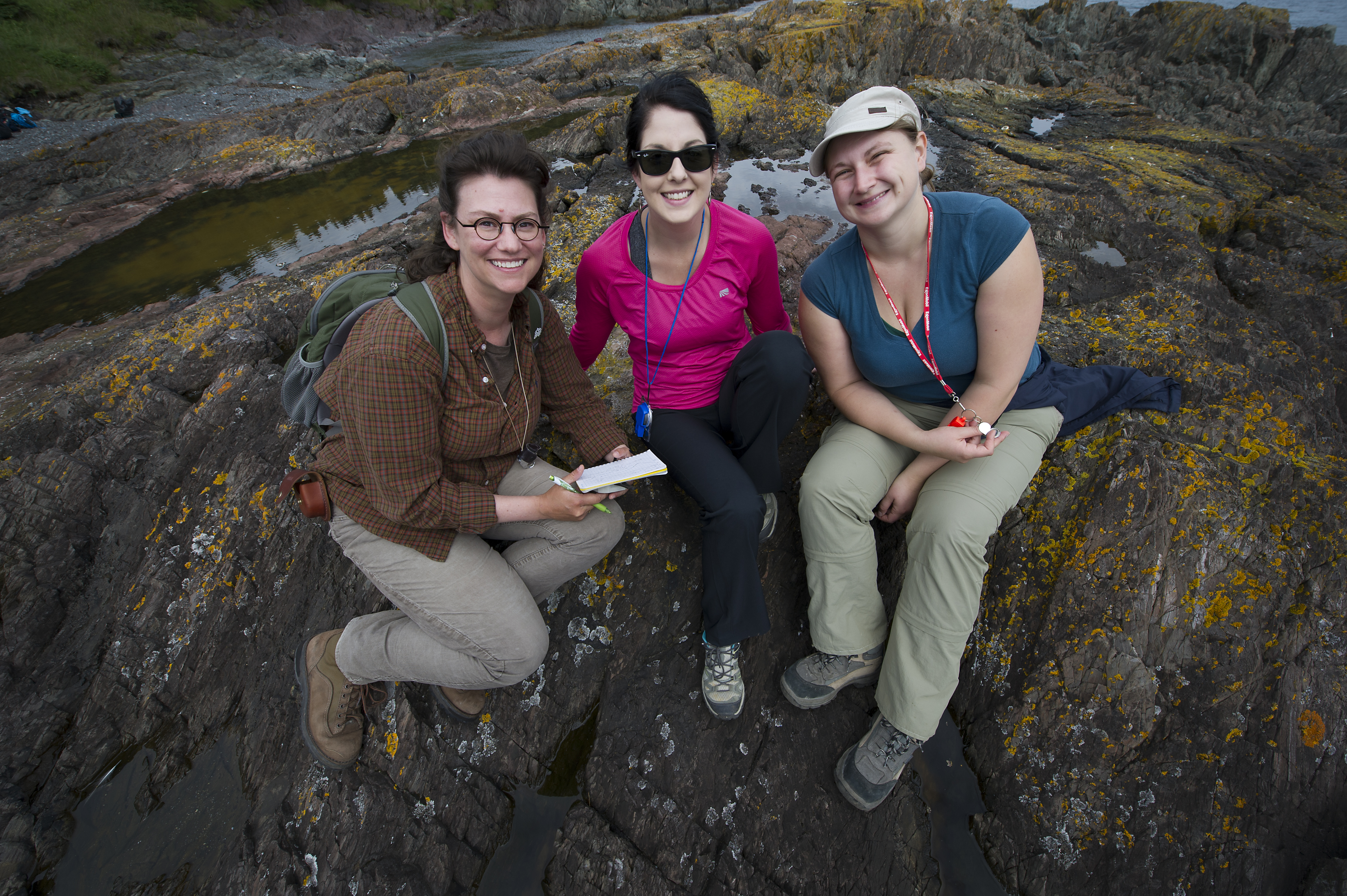 Three students in the field sitting on the bedrock with a compass in hand